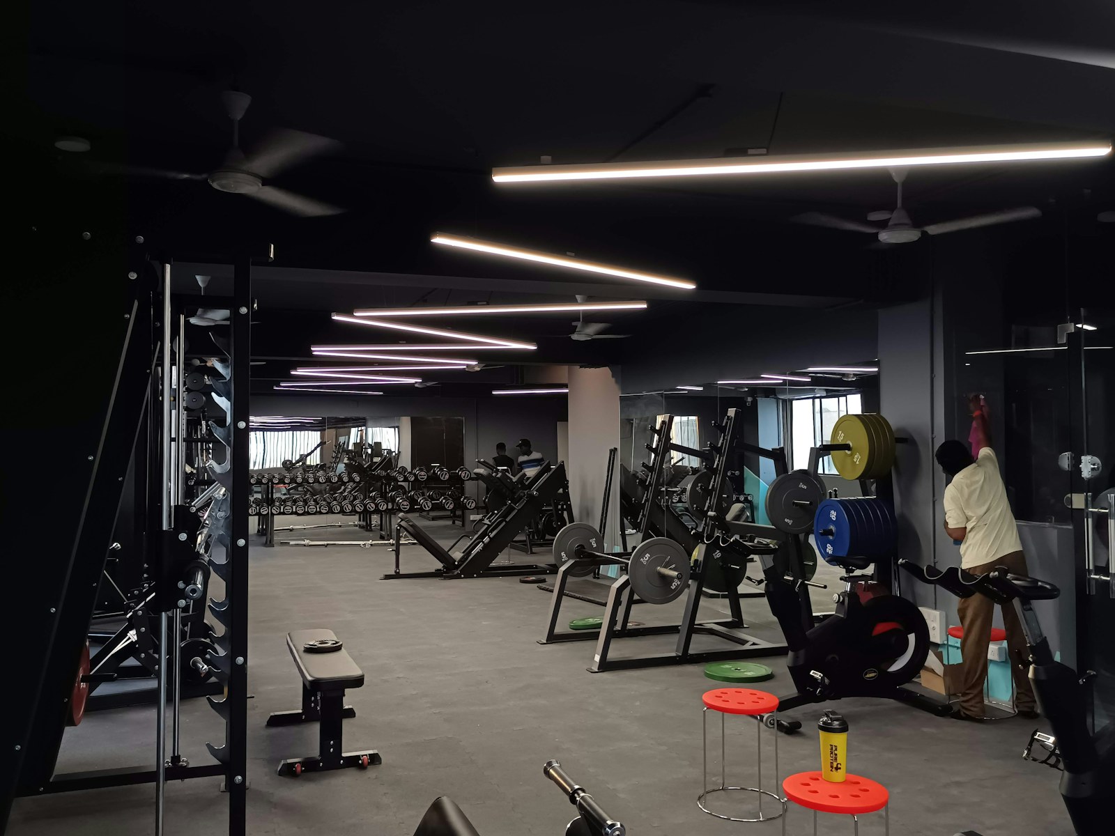 A dimly lit gym interior lined with weight machines, benches and loaded barbells, with a person adjusting equipment near the wall.