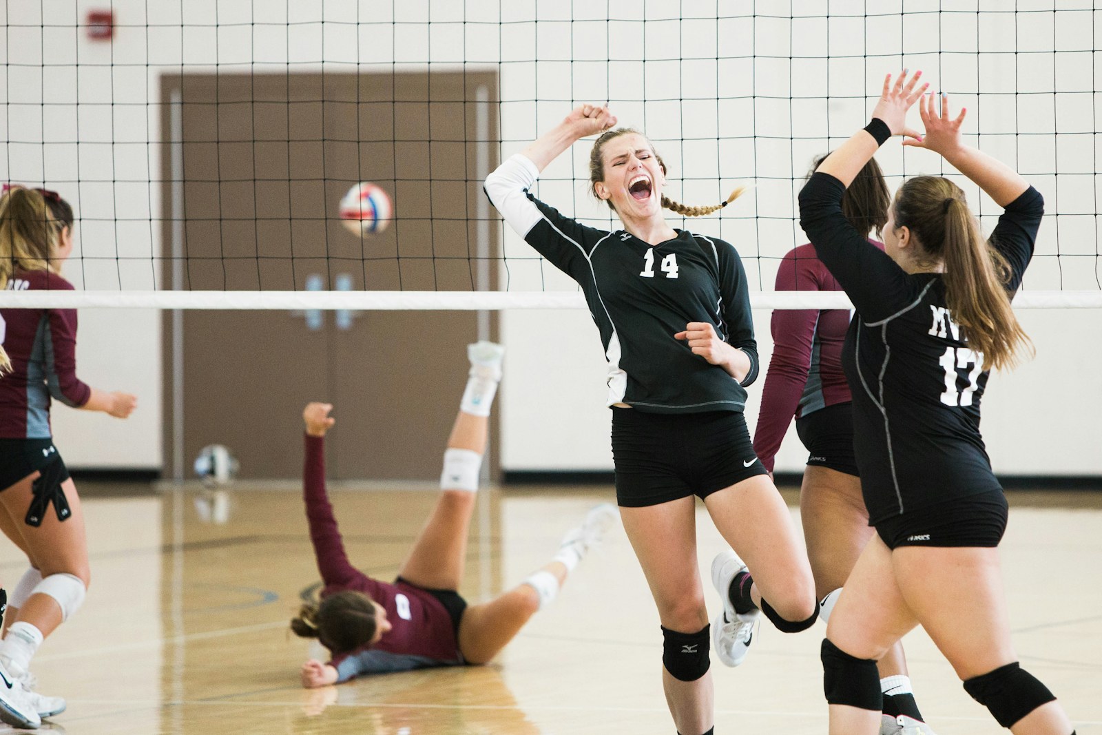 Women's volleyball players celebrate a point at the net on an indoor court as the ball flies past in the background.