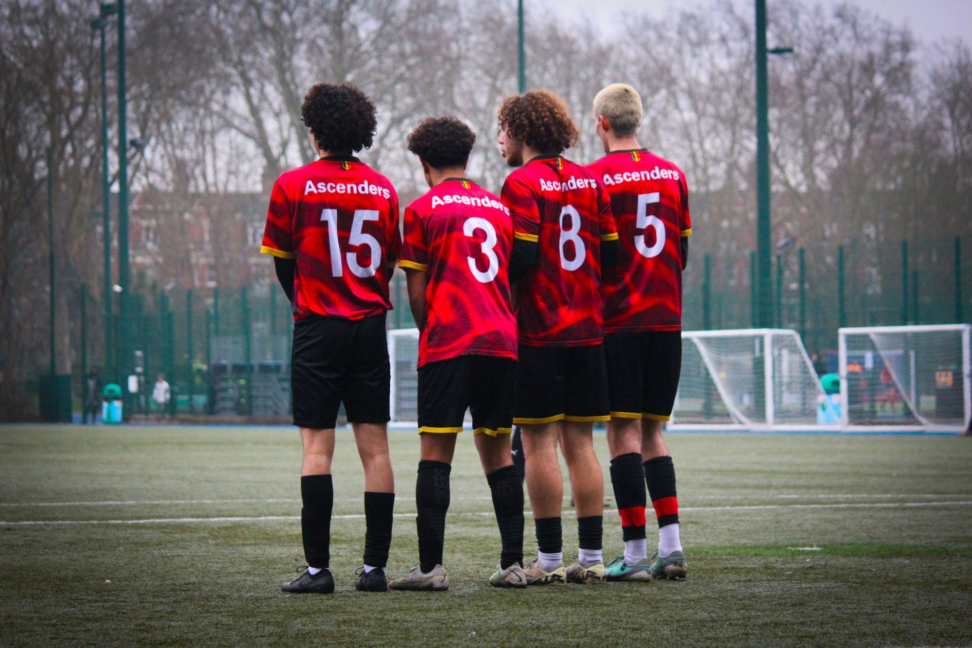 Four footballers seen from behind, standing shoulder to shoulder in matching red and black numbered jerseys, lined up together on a grass pitch under an overcast sky.